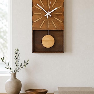 Puede incluir: Un reloj de pared rectangular de madera marr&oacute;n oscuro con una esfera estilo rayos de sol que presenta marcadores de hora de madera clara. Tiene manecillas blancas y un p&eacute;ndulo circular de madera clara que oscila en un compartimento abierto.