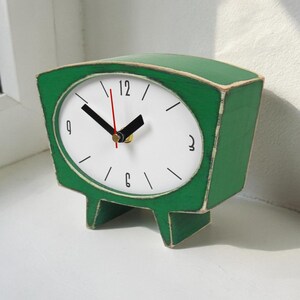May include: A green wooden table clock with a white face, black hour and minute hands, and a thin red second hand. The clock features a wide, curved top, a tapered base with two feet, and black numerals at the 12, 3, 6, and 9 o'clock positions.