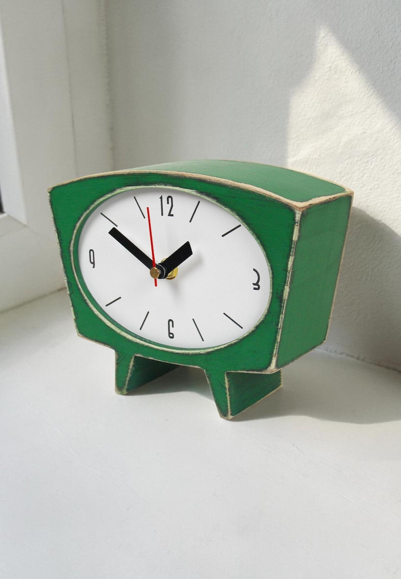 May include: A green wooden table clock with a white face, black hour and minute hands, and a thin red second hand. The clock features a wide, curved top, a tapered base with two feet, and black numerals at the 12, 3, 6, and 9 o'clock positions.
