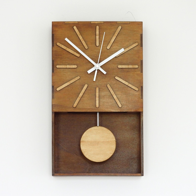 Puede incluir: Un reloj de pared rectangular de madera con acabado marr&oacute;n oscuro y doce marcadores de hora de madera clara dispuestos en c&iacute;rculo. El reloj tiene manecillas blancas para las horas y los minutos y un p&eacute;ndulo circular de madera suspendido.