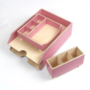 Op de afbeelding: Een set roze houten bureau-organizers met natuurlijke houten interieurs. De set bevat een grote lade, een kleinere lade met vakken en een rechthoekige doos met drie secties. Voor op kantoor of thuis.