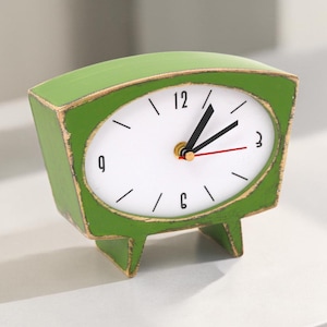 May include: A green, retro-style tabletop clock with a white oval clock face. The clock has black numerals, black hands, and a red second hand. The clock is approximately 5 inches tall.