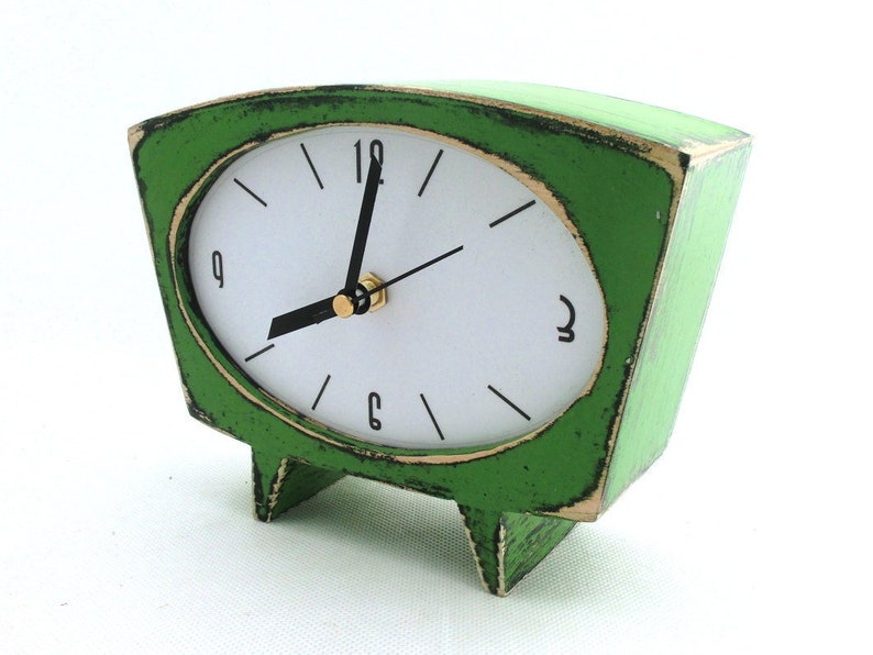 May include: A trapezoidal table clock with a distressed green finish and a white face. The clock features black hour markers, black hands, and numerals at the 12, 3, 6, and 9 o'clock positions. It stands on two small, integrated green feet.