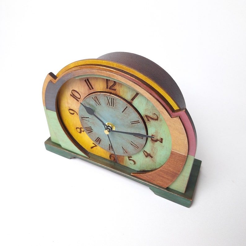 May include: A wooden mantel clock with a multi-coloured design. The clock face is a light blue with Roman numerals and gold hands. The clock has a dark brown top and a green base. The clock measures approximately 20 cm wide and 15 cm tall.