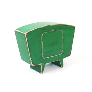 May include: A green wooden box with a curved top and a rectangular panel on the front. The edges of the box and the panel are highlighted with a distressed white finish. It stands on two small, integrated wooden feet and has a single brass screw.