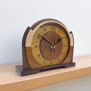 May include: A wooden mantel clock with a layered design in shades of brown. The clock face features black hands and numerals. The clock sits on a rectangular base and is displayed on a wooden shelf. The clock is approximately 20 cm in height.