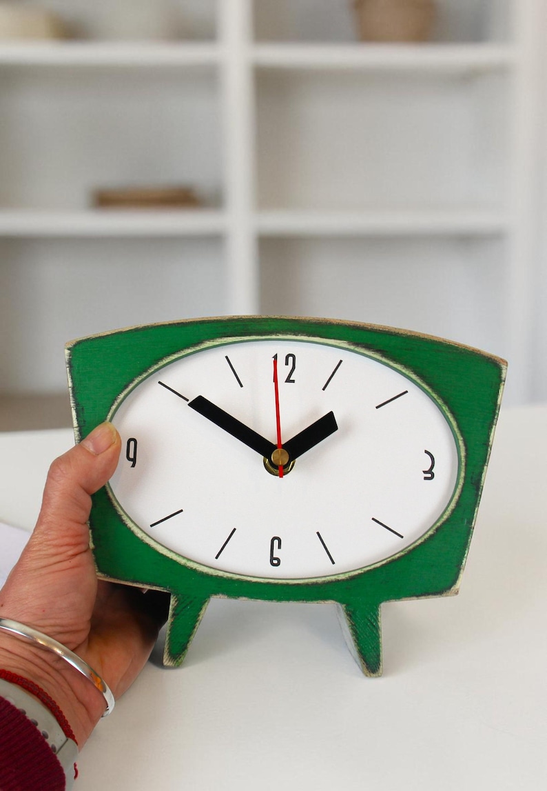 May include: A green wooden table clock with a white oval face, black hour and minute hands, and a thin red second hand. The clock features black numerals at the 12, 3, 6, and 9 positions, with simple black lines for the remaining hour markers.