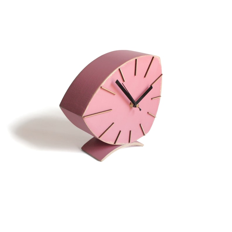 Peut inclure: Une horloge en bois rose avec un cadran en forme de feuille et des aiguilles noires. L'horloge est sur un socle incurv&eacute;.