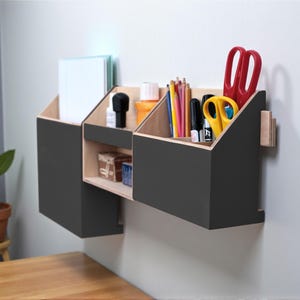 Op de afbeelding: Een moderne wandorganizer. Het heeft drie compartimenten: twee schuine secties en een kleine plank. De organizer is donkergrijs met een natuurlijk houten interieur. Het bevat briefpapier zoals potloden, scharen en notitieboekjes. Afmetingen in centimeters.