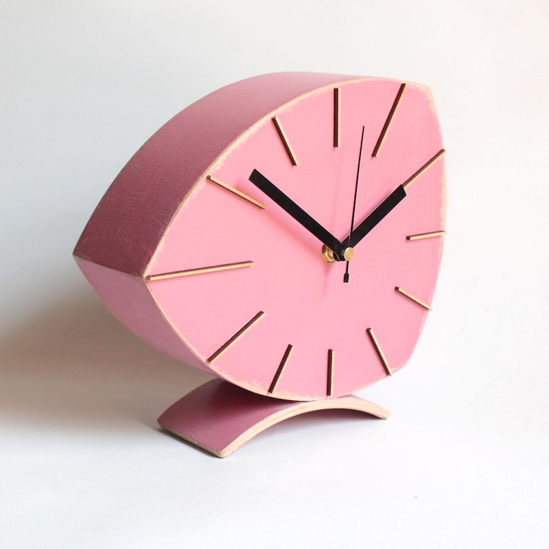 Peut inclure: Une horloge en bois rose avec des aiguilles noires et des accents dor&eacute;s. L'horloge est en forme de feuille et a une base incurv&eacute;e.