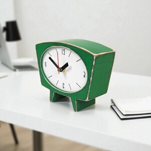 May include: A green table clock with a trapezoidal shape and a white face. The clock features black hour and minute hands, a red second hand, and black numerals at the 12, 3, 6, and 9 positions. The clock stands on two short, wide legs.