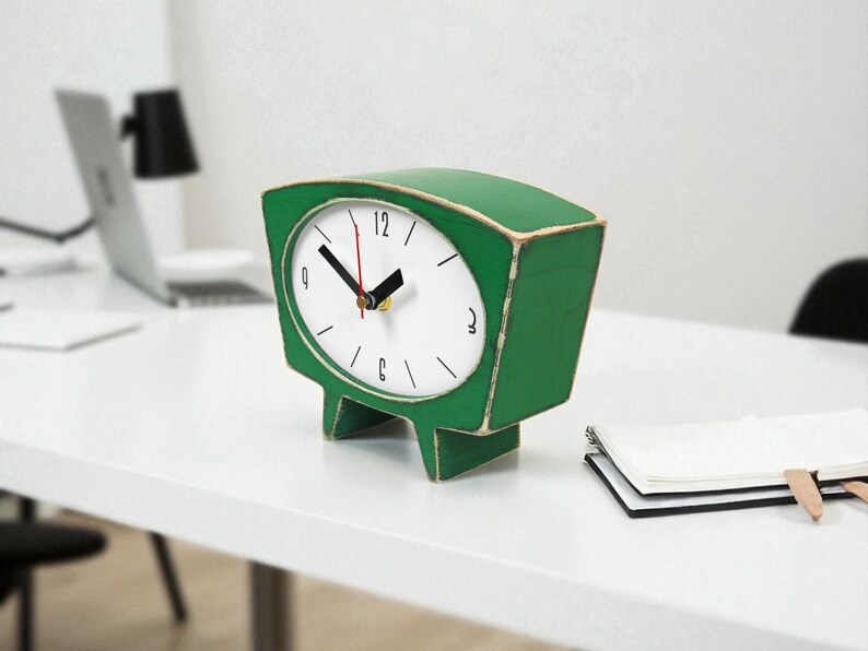 May include: A green table clock with a trapezoidal shape and a white face. The clock features black hour and minute hands, a red second hand, and black numerals at the 12, 3, 6, and 9 positions. The clock stands on two short, wide legs.