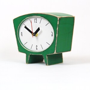 May include: A green wooden table clock with a white circular face, black hour and minute hands, and a thin red second hand. The clock features black numerals at the 12, 3, 6, and 9 positions, with simple black lines for the remaining hours.