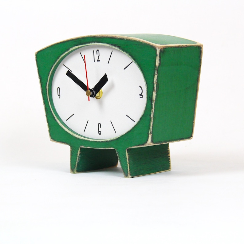 May include: A green wooden table clock with a white circular face, black hour and minute hands, and a thin red second hand. The clock features black numerals at the 12, 3, 6, and 9 positions, with simple black lines for the remaining hours.