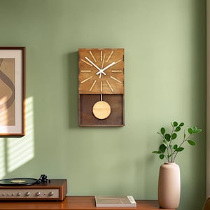 Puede incluir: Un reloj de pared rectangular de madera con marco marr&oacute;n oscuro, esfera de madera clara con manecillas blancas y un p&eacute;ndulo circular que oscila en un compartimento inferior abierto. El reloj tiene doce marcas lineales.