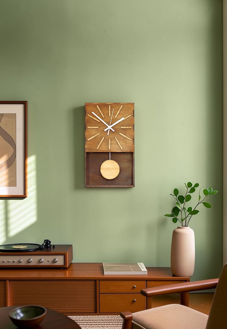 Puede incluir: Un reloj de pared rectangular de madera con marco marr&oacute;n oscuro, esfera de madera clara con manecillas blancas y un p&eacute;ndulo circular que oscila en un compartimento inferior abierto. El reloj tiene doce marcas lineales.