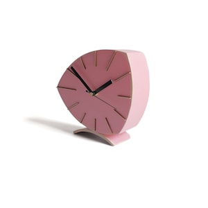 Peut inclure: Une horloge de forme triangulaire rose avec des accents dor&eacute;s et des aiguilles noires. L'horloge est sur une surface blanche.