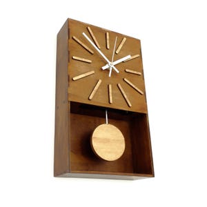Puede incluir: Un reloj de pared rectangular de madera con acabado marr&oacute;n oscuro. La esfera presenta doce marcadores de hora de madera clara y dos manecillas blancas. Un p&eacute;ndulo circular de madera cuelga de una varilla plateada en la secci&oacute;n inferior abierta.