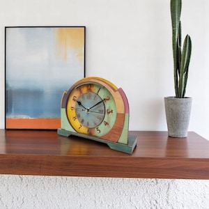 May include: A colourful wooden mantel clock with a vintage design. The clock face is light blue with black hands and Roman numerals. The clock's frame features a rainbow of colours, including yellow, green, and pink. The clock is approximately 25 cm wide.