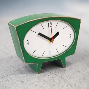May include: A green tabletop clock with a trapezoidal wooden frame featuring distressed edges. The white clock face displays black numerals at 3, 6, 9, and 12, with black hour and minute hands and a thin red second hand. The clock stands on two feet.