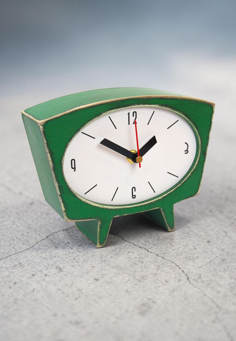 May include: A green tabletop clock with a trapezoidal wooden frame featuring distressed edges. The white clock face displays black numerals at 3, 6, 9, and 12, with black hour and minute hands and a thin red second hand. The clock stands on two feet.