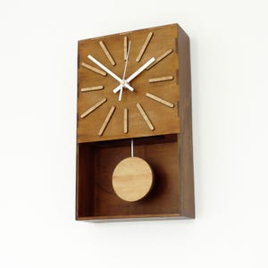 Puede incluir: Un reloj de pared rectangular hecho de madera marr&oacute;n con una cara plana que presenta doce marcadores de hora de madera en relieve. El reloj tiene dos manecillas blancas y un p&eacute;ndulo circular de madera suspendido en la secci&oacute;n inferior.