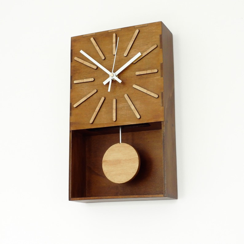 Puede incluir: Un reloj de pared rectangular hecho de madera marr&oacute;n con una cara plana que presenta doce marcadores de hora de madera en relieve. El reloj tiene dos manecillas blancas y un p&eacute;ndulo circular de madera suspendido en la secci&oacute;n inferior.