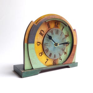 May include: A wooden mantel clock with a multi-coloured design. The clock face is a faded blue with black hands and Roman numerals. The clock's frame features shades of green, yellow, and brown. The clock sits on a green base.