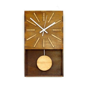 Puede incluir: Un reloj de pared rectangular hecho de madera marr&oacute;n oscura con un acabado de madera m&aacute;s clara en el p&eacute;ndulo circular y los marcadores de hora. La esfera tiene manecillas blancas y doce marcadores rectangulares espaciados.