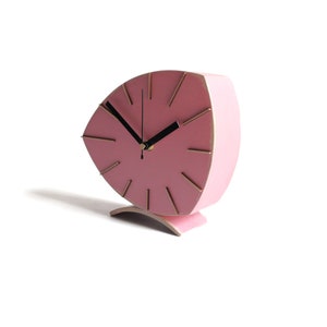 Peut inclure: Une horloge rose en forme de feuille avec des marqueurs d'heures dor&eacute;s et des aiguilles noires. L'horloge est sur un fond blanc.