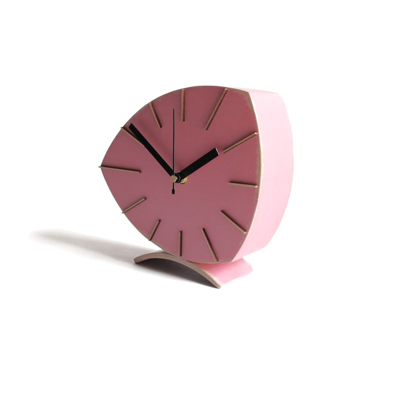 Peut inclure: Une horloge rose en forme de feuille avec des marqueurs d'heures dor&eacute;s et des aiguilles noires. L'horloge est sur un fond blanc.