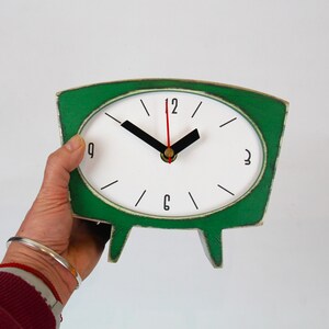 May include: A tabletop clock with a green, trapezoidal wooden frame and a white oval face. The clock features black hour and minute hands, a thin red second hand, and black numerals at the 12, 3, 6, and 9 o'clock positions with simple lines.