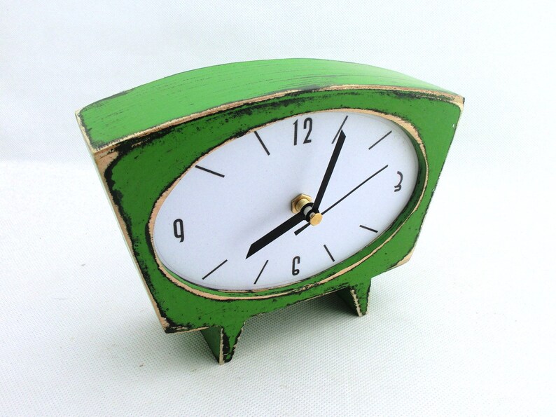 May include: A trapezoidal table clock with a distressed bright green finish and black edges. The clock face is white with black numerals at 12, 3, 6, and 9, featuring black hour and minute hands and a gold-toned center pin mechanism.