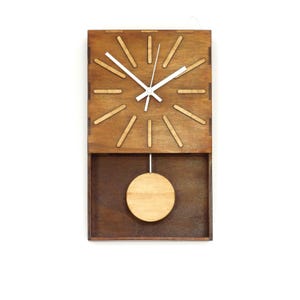 Puede incluir: Un reloj de pared rectangular hecho de madera te&ntilde;ida de marr&oacute;n oscuro. La esfera presenta marcadores de hora de madera clara y manecillas blancas. Debajo, un p&eacute;ndulo circular de madera clara cuelga de una fina varilla met&aacute;lica.