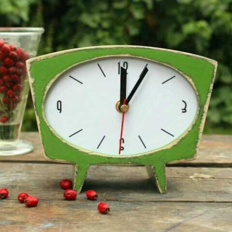 May include: A green table clock with a white oval face, black hour and minute hands, and a thin red second hand. The clock features black numerals at the twelve, three, six, and nine o'clock positions, set within a distressed green frame.