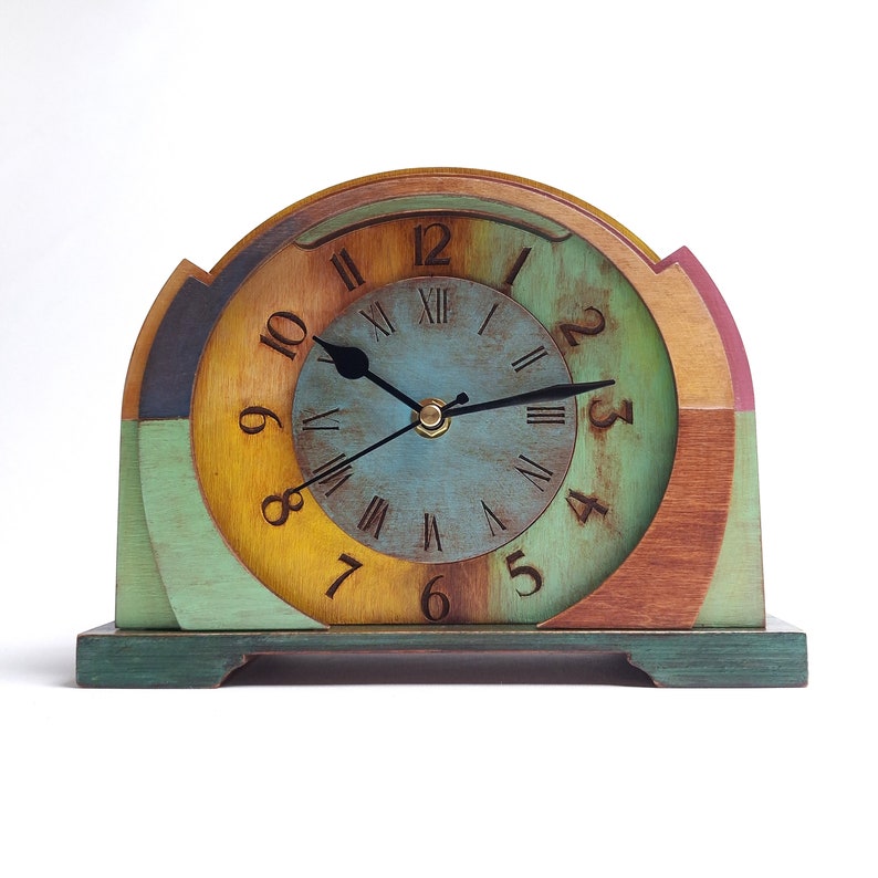 May include: A colourful wooden clock with an arched design. The clock face is faded blue with Roman numerals and black hands. The frame features green, yellow, brown, and pink. The clock sits on a green base. A decorative item.