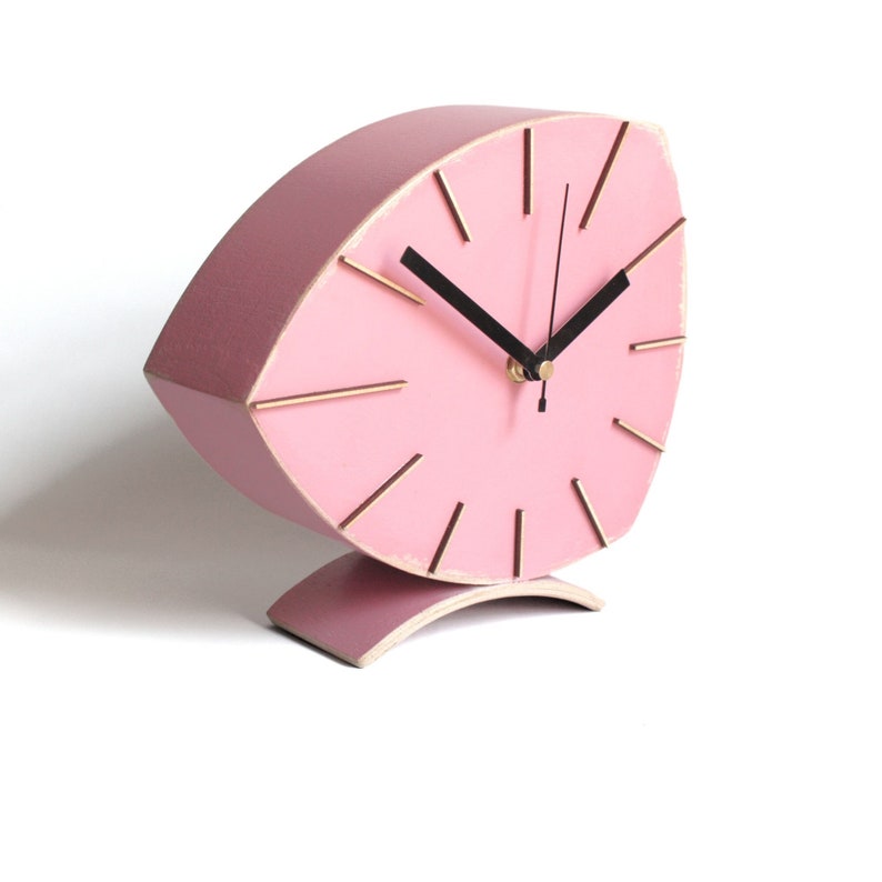 Peut inclure: Une horloge en bois rose avec des accents dor&eacute;s et des aiguilles noires. L'horloge est en forme de feuille et a une base incurv&eacute;e.