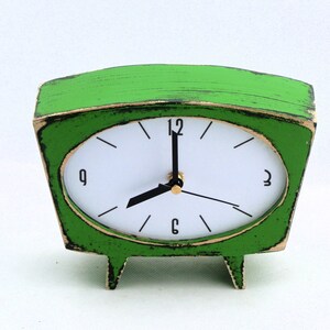 May include: A bright green table clock with a trapezoidal frame and a distressed finish. The clock face is white with black numerals at 12, 3, 6, and 9, featuring black hour and minute hands and a thin black second hand on a gold center.