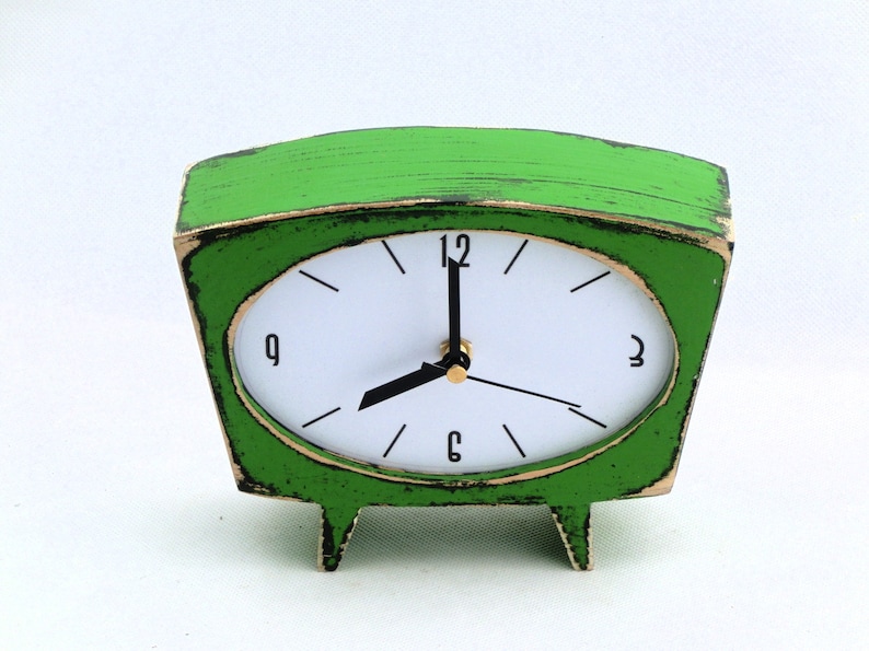 May include: A bright green table clock with a trapezoidal frame and a distressed finish. The clock face is white with black numerals at 12, 3, 6, and 9, featuring black hour and minute hands and a thin black second hand on a gold center.