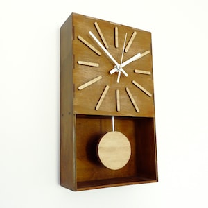 Puede incluir: Un reloj de pared rectangular de madera con acabado marr&oacute;n medio. La esfera presenta doce marcadores de madera clara y manecillas blancas finas. Debajo de la esfera, un p&eacute;ndulo circular de madera cuelga de una varilla blanca fina.