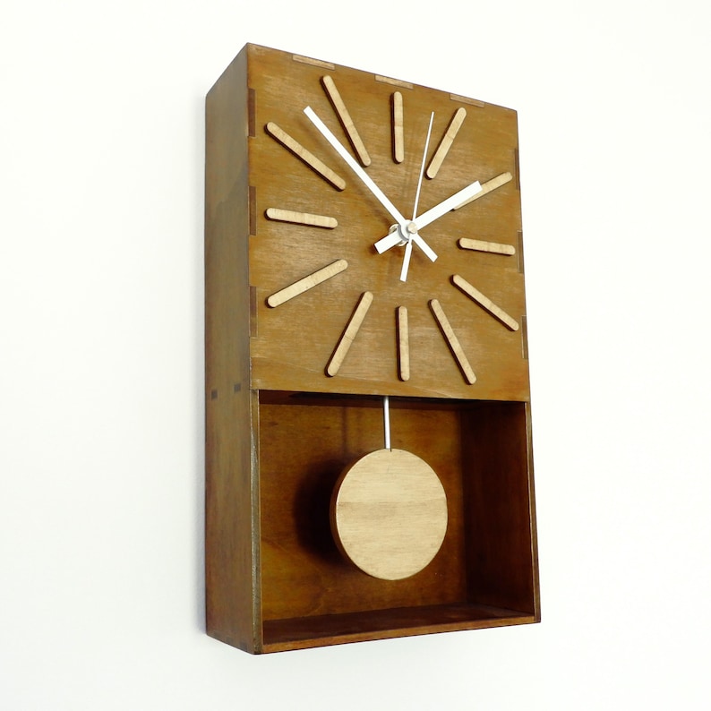 Puede incluir: Un reloj de pared rectangular de madera con acabado marr&oacute;n medio. La esfera presenta doce marcadores de madera clara y manecillas blancas finas. Debajo de la esfera, un p&eacute;ndulo circular de madera cuelga de una varilla blanca fina.