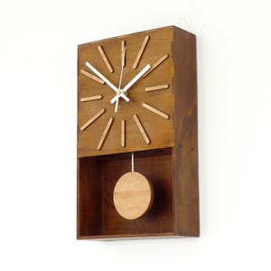 Puede incluir: Un reloj de pared rectangular de madera con acabado marr&oacute;n oscuro. La esfera presenta doce marcadores de madera marr&oacute;n claro y manecillas blancas para horas, minutos y segundos. Un p&eacute;ndulo circular de madera cuelga en un compartimento.