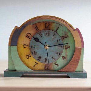 May include: A colourful wooden clock with a round face and a semi-circular top. The clock face is blue with black hands and Roman numerals. The clock is painted in shades of green, yellow, brown, and purple. The clock measures approximately 18 cm wide and 20 cm tall.