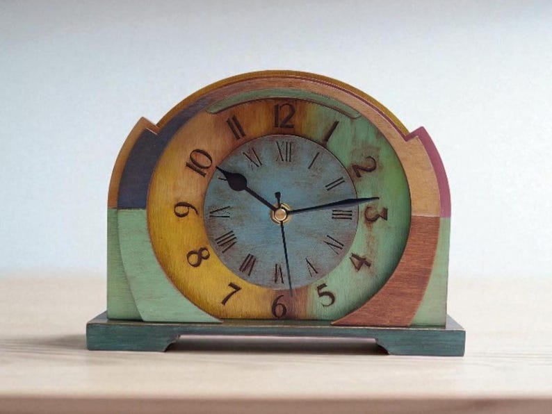May include: A colourful wooden clock with a round face and a semi-circular top. The clock face is blue with black hands and Roman numerals. The clock is painted in shades of green, yellow, brown, and purple. The clock measures approximately 18 cm wide and 20 cm tall.