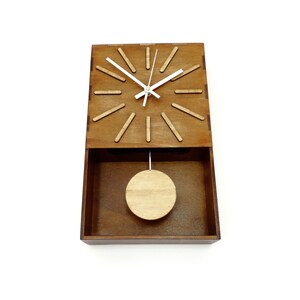 Puede incluir: Un reloj de pared rectangular de madera con acabado marr&oacute;n oscuro. La esfera presenta doce marcadores de hora de madera clara, manecillas blancas y un p&eacute;ndulo circular suspendido en la secci&oacute;n inferior abierta del marco de madera.