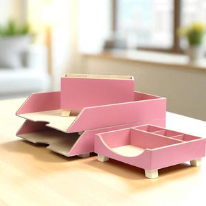 Op de afbeelding: Een set roze houten bureau-organizers. De set bevat een papierbak met twee niveaus, een pennenhouder en een klein dienblad met vakken. De organizers hebben een licht houten interieur.