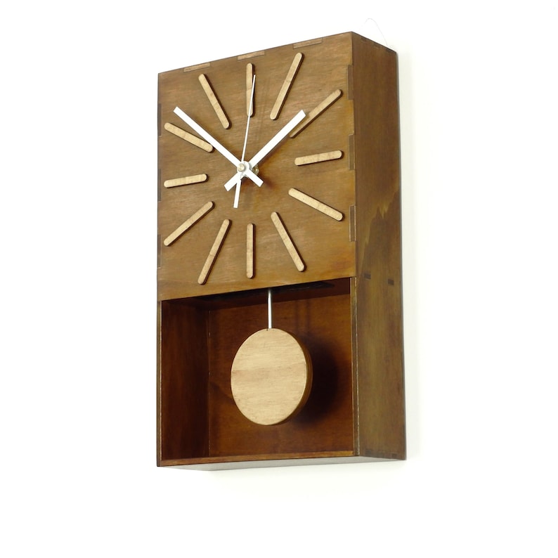 Puede incluir: Un reloj de pared rectangular hecho de madera marr&oacute;n oscura con acabado natural. La esfera presenta doce marcadores de hora en forma de palitos de madera clara, manecillas de metal blanco y un p&eacute;ndulo circular de madera en la parte inferior.