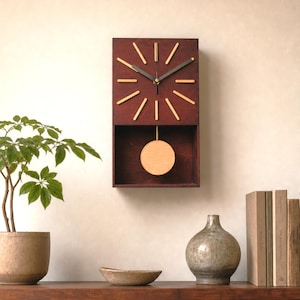 Può includere: Un orologio da parete rettangolare in legno marrone scuro. Il quadrante presenta indicatori delle ore a bastoncino color oro e lancette nere. Un pendolo in legno è appeso sotto. Il design è minimalista.