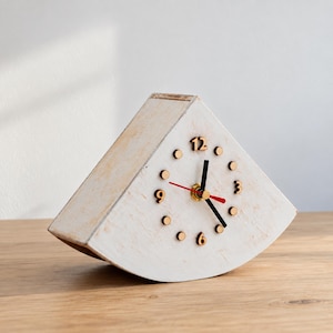 Può includere: Un orologio da tavolo in legno di forma triangolare con finitura bianca anticata. Il quadrante presenta numeri marrone scuro per 3, 6, 9 e 12, con piccoli indicatori circolari per le altre ore. Ha lancette nere e una lancetta dei secondi rossa.