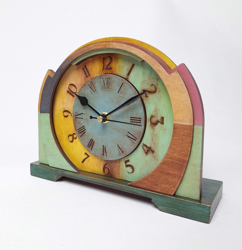 May include: A wooden mantel clock with a multi-coloured face and a green base. The clock face has Roman numerals and black hands. The clock is painted in shades of blue, yellow, green, brown, and pink. The clock measures approximately 25 cm in height.